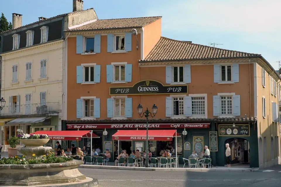 Une place de Dieulefit bordée d'immeubles aux volets bleus, avec des terrasses de café sous des stores rouges et une fontaine au premier plan