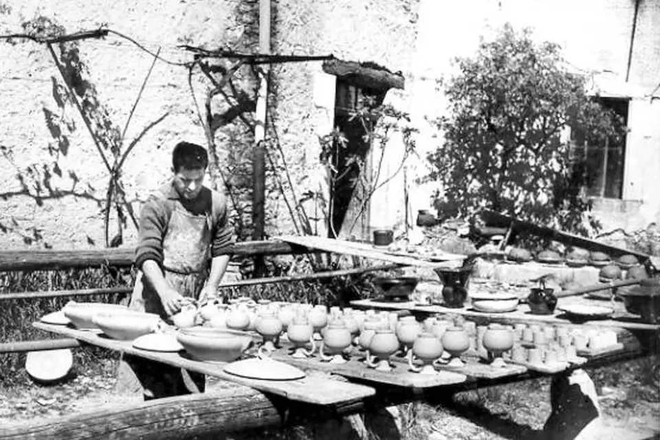 Photographie ancienne d'un potier de Dieulefit debout derrière de longues tables de séchage couvertes de plats, de pots et de jattes, devant un atelier